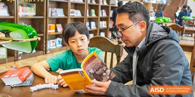 A male teacher from Dingwen Academy Hangzhou reading a book aloud to a young student in a library or reading corner.