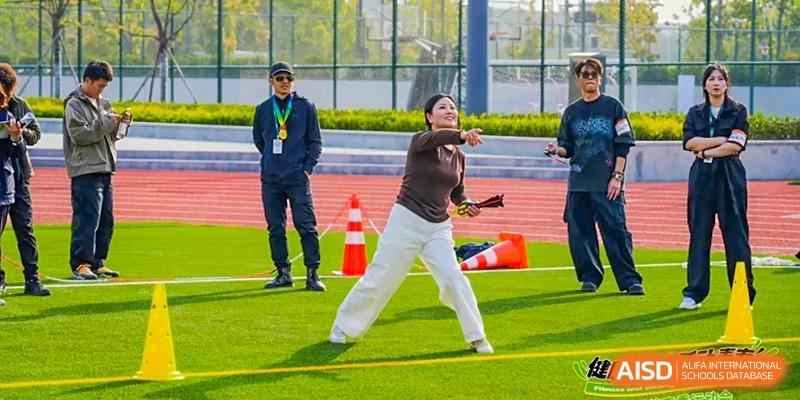 A student from Dingwen Academy Hangzhou performing a throwing action (like shot put or javelin) on a sports field while instructors watch.
