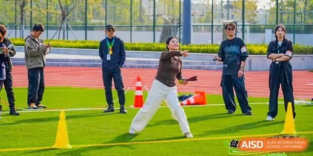 A student from Dingwen Academy Hangzhou performing a throwing action (like shot put or javelin) on a sports field while instructors watch.