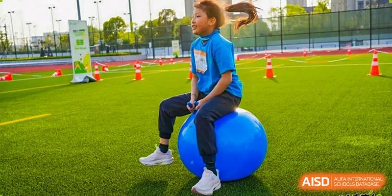 A female student from Dingwen Academy Hangzhou bouncing on a large blue exercise ball on a turf field marked with orange cones.