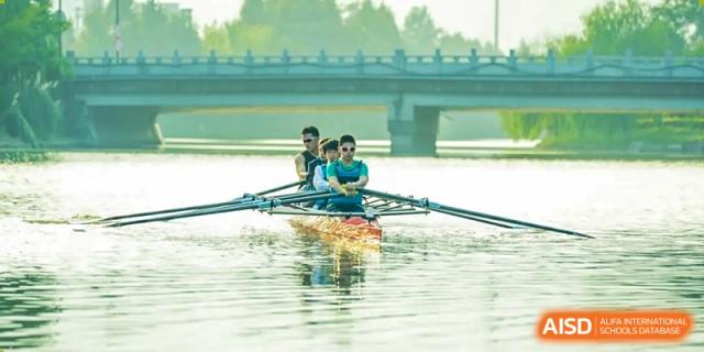 Students from Dingwen Academy Hangzhou rowing a sculling boat on the water beneath a bridge during a practice session.