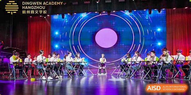 Students and faculty from Dingwen Academy Hangzhou seated on a stage during an evening ceremony with a large circular screen backdrop.