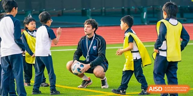 A coach from Dingwen Academy Hangzhou kneeling to speak with two young students while holding a soccer ball on a turf field.