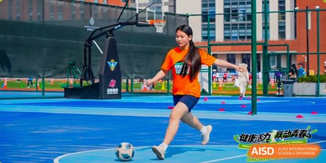 A female student from Dingwen Academy Hangzhou wearing an orange shirt and shorts, kicking a soccer ball on an outdoor court with a basketball hoop visible.