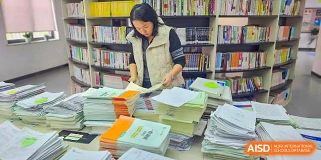 A student from Henan Zhengzhou Jianye International School organizing a large stack of textbooks and papers in the school library or resource area.
