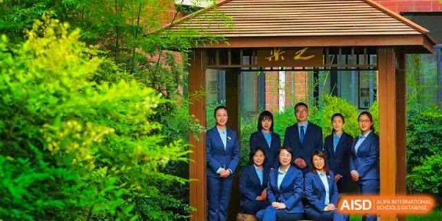 A small group of students and a teacher from Henan Zhengzhou Jianye International School standing together in a green, outdoor pavilion area.