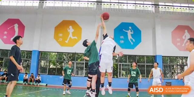 Students from Henan Zhengzhou Jianye International School competing in an indoor basketball game, with one player jumping up to shoot or block the ball.