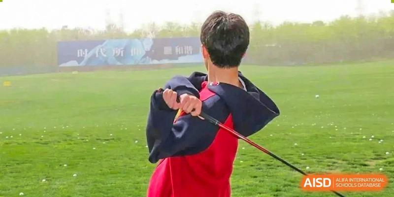 A student from Henan Zhengzhou Jianye International School practicing a golf swing outdoors on a green course, viewed from behind.