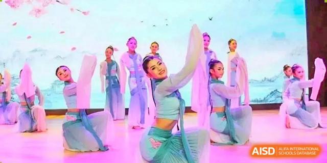 A group of female students from Henan Zhengzhou Jianye International School performing a traditional or cultural dance in light blue and white costumes on stage.