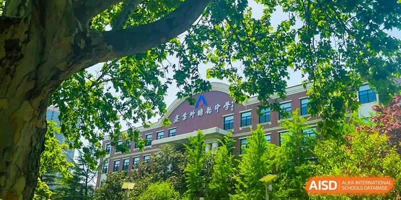 The main building of Henan Zhengzhou Jianye International School, visible through the lush green trees on a sunny day.