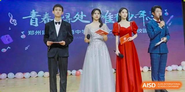 Students from Henan Zhengzhou Jianye International School formally dressed, standing on stage with certificates or awards, including a female student in a white gown.