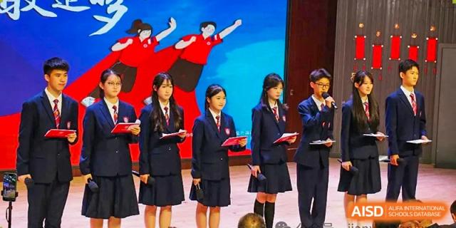 Students from Henan Zhengzhou Jianye International School in dark uniforms standing on stage during a ceremony, with performers in red visible in the background.
