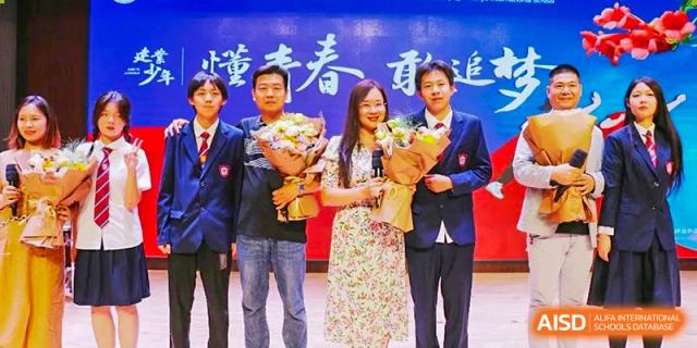 A group of students and faculty members from Henan Zhengzhou Jianye International School holding bouquets of flowers while posing on stage at an event.