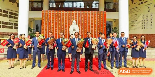 A group of teachers or staff members from Henan Zhengzhou Jianye International School posing with awards on a red carpet inside a school hall with a traditional statue.