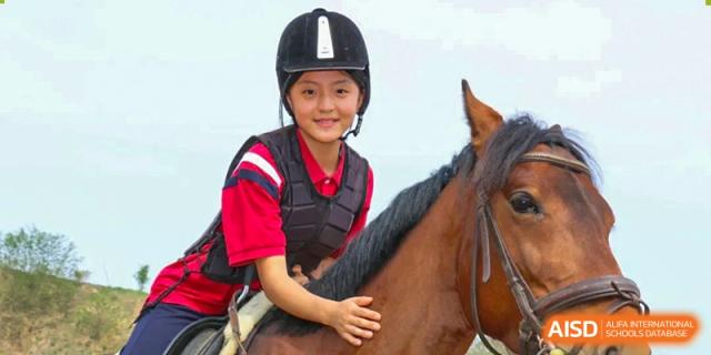 A female student from Henan Zhengzhou Jianye International School wearing safety gear, smiling while riding a brown horse during an equestrian activity.