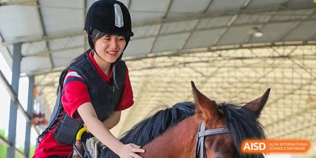 A student from Henan Zhengzhou Jianye International School petting or sitting on the neck of a brown horse during an equestrian training session.