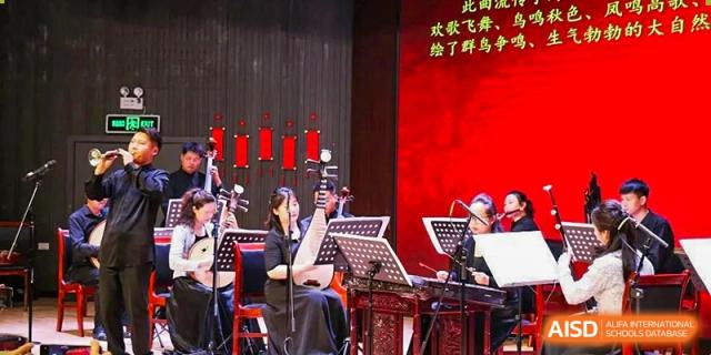 Students from Henan Zhengzhou Jianye International School performing traditional Chinese music with various instruments on a stage with red backlighting.