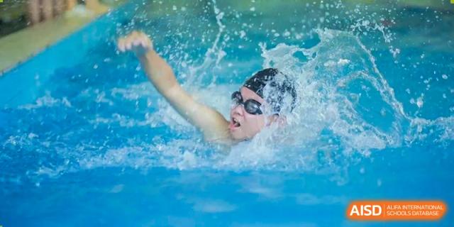 A student from Henan Zhengzhou Jianye International School wearing goggles and a swim cap, swimming vigorously in a blue pool.