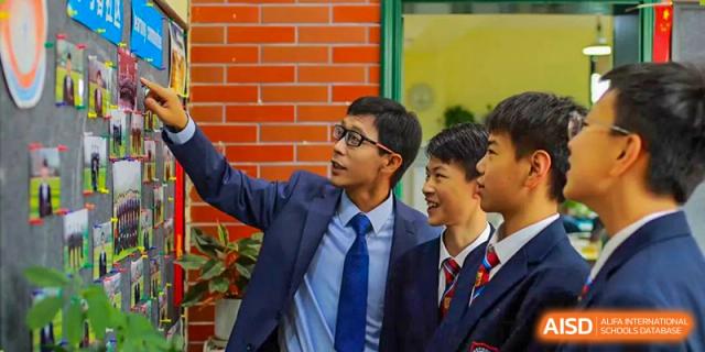 A male teacher from Henan Zhengzhou Jianye International School in a suit interacting with two students in uniform against a red brick wall background.