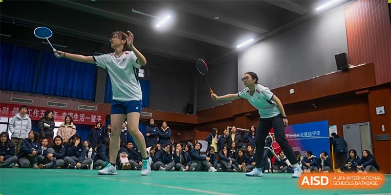 Students from International Department of Jinling High School Hexi Campus playing badminton in an indoor gymnasium while others watch from the sidelines.