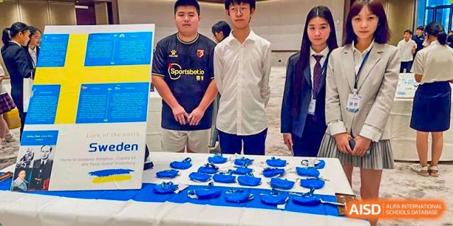 Three students from International Department of Jinling High School Hexi Campus standing behind a table displaying blue model boats or items, possibly at a competition or fair.