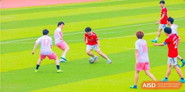 Two students from International Department of Jinling High School Hexi Campus playing soccer or practicing on a bright green field, with one student dribbling the ball.