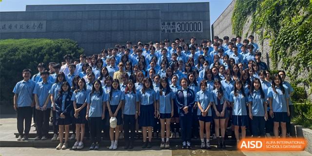 A large group of students from International Department of Jinling High School Hexi Campus, dressed in matching blue shirts and skirts/trousers, posing for a group photo outdoors in front of a building wall.