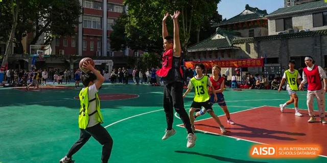 Students from the Guangdong Overseas Chinese High School International Curriculum playing an outdoor game of basketball, with one student leaping to shoot or block a ball.