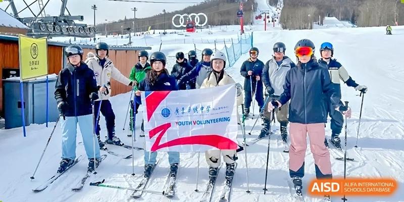Guangdong Overseas Chinese High School International Curriculum students on a ski trip or winter event, holding a "Youth Volunteering" banner on a snowy slope.