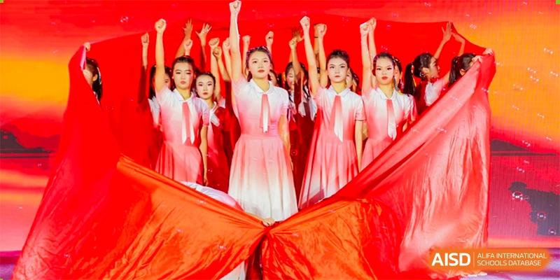 A group of female students from the Guangdong Overseas Chinese High School International Curriculum performing a vibrant dance in pink and white costumes on a stage.