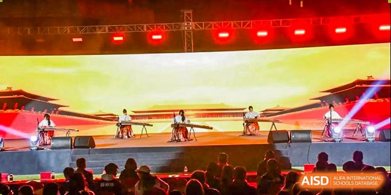 A musical performance at the Guangdong Overseas Chinese High School International Curriculum, with musicians playing traditional instruments on a stage with red lighting.