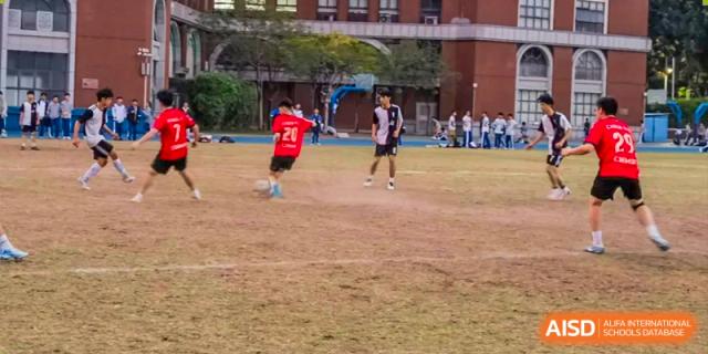 Guangdong Overseas Chinese High School International Curriculum students playing an intense game of soccer/football on a dirt or dry field.