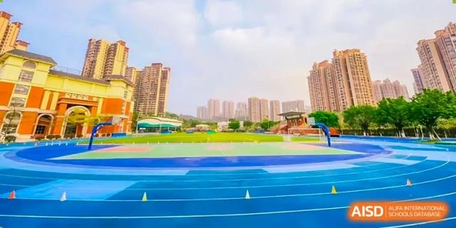The track and field facilities of the Guangdong Overseas Chinese High School International Curriculum, featuring a bright blue running track and green field, with city buildings in the background.
