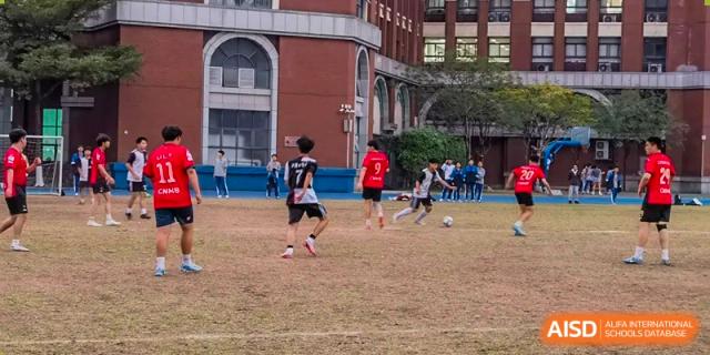 Students from the Guangdong Overseas Chinese High School International Curriculum playing a game of soccer/football on a grass field near the school buildings.