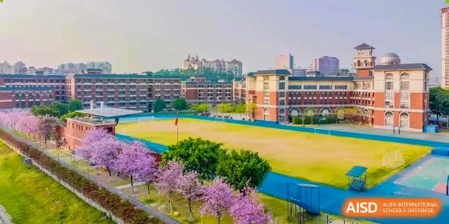 The sports field and main campus buildings of the Guangdong Overseas Chinese High School International Curriculum, with blossoming purple trees in the foreground.