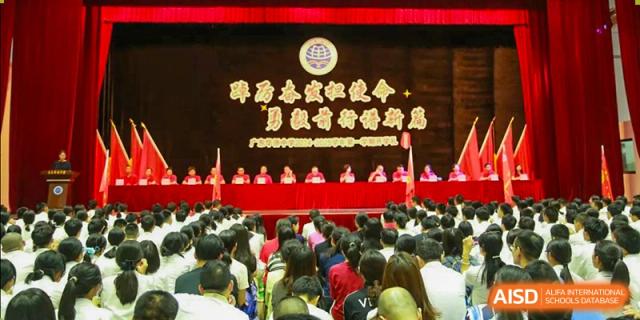 A large formal assembly or conference held indoors at the Guangdong Overseas Chinese High School International Curriculum, with a panel of speakers and many attendees.