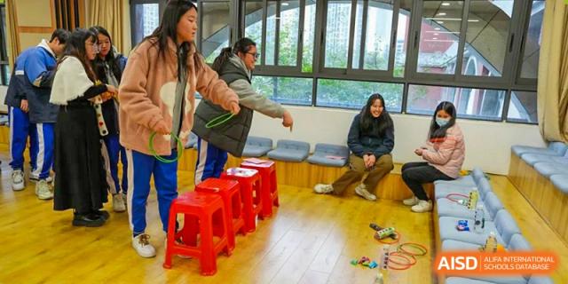Students from the Guangdong Overseas Chinese High School International Curriculum playing a team game or activity indoors with red stools.