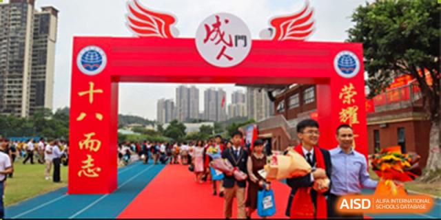 Students from the Guangdong Overseas Chinese High School International Curriculum entering through a large archway/gate during an outdoor event, possibly a marathon or race.