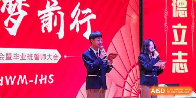 A Wuhan Weiming Senior High School student speaking on a microphone on a stage with a red background during a school event or ceremony.