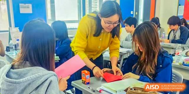 A female teacher in a yellow sweater bending over to help a student with a red folder at the Shanghai Jincai High School International Division.