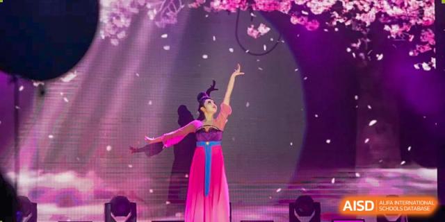 A female student in a traditional pink and purple costume dancing on an indoor stage at the Shanghai Jincai High School International Division.