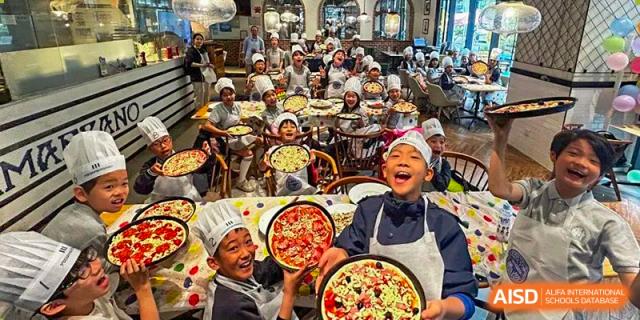A large group of students in chef's hats laughing and holding up pizzas in a dining or cooking area at the Shanghai Jincai High School International Division.