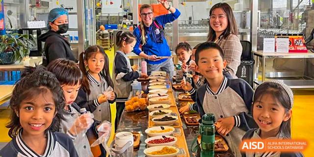 A teacher smiling with a group of students at a long table with various food items during a cooking or nutrition class at the Shanghai Jincai High School International Division.