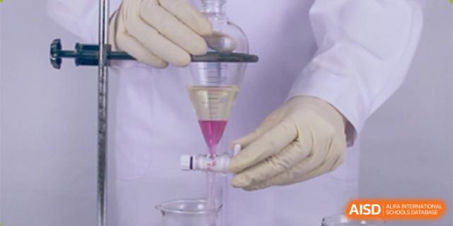 A pair of gloved hands performing a liquid-liquid separation using a small separatory funnel, dripping pink liquid into a test tube. Canada British Colombia International Schools-Hefei.