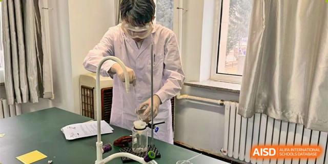 A male student in a lab coat and safety glasses carefully conducting a chemistry experiment at a lab bench near a window. Canada British Colombia International Schools-Hefei.