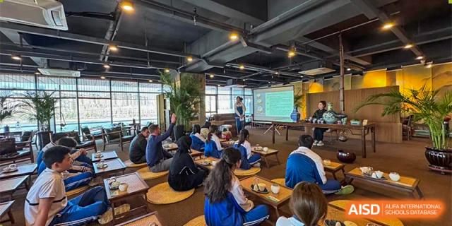 Students seated on floor cushions in a casual, cafe-like setting for a class or presentation, with an instructor standing near a projector screen. Canada British Colombia International Schools-Hefei.