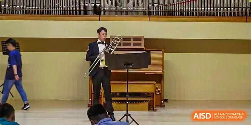 A student dressed in formal wear playing the trombone during a musical performance indoors. Canada British Colombia International Schools-Hefei.