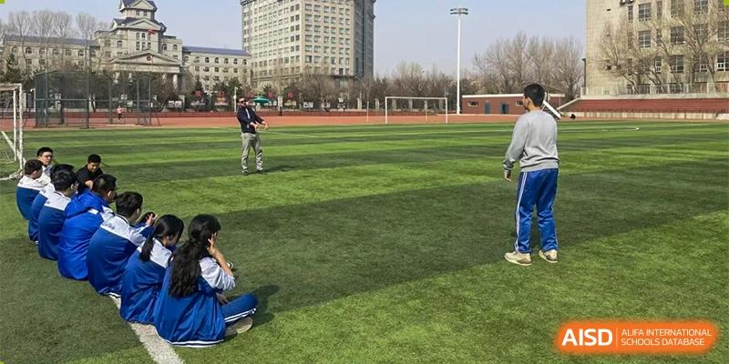 Students sitting and standing on a large green sports field near a tall school building, perhaps during an outdoor class or break. Canada British Colombia International Schools-Hefei.