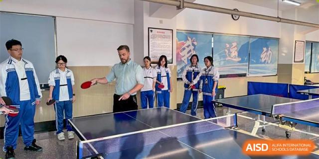 A teacher instructing or playing table tennis (ping pong) with students in an indoor school area. Canada British Colombia International Schools-Hefei.