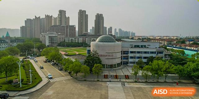 An aerial view of the school campus buildings, including a prominent circular dome structure, set against the city skyline of Wuhan Foreign Languages School International Division.